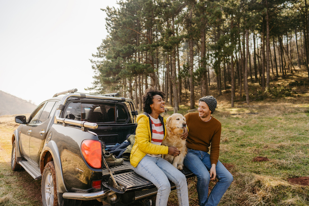 A couple sits on the gate of their pickup truck, petting their dog. 