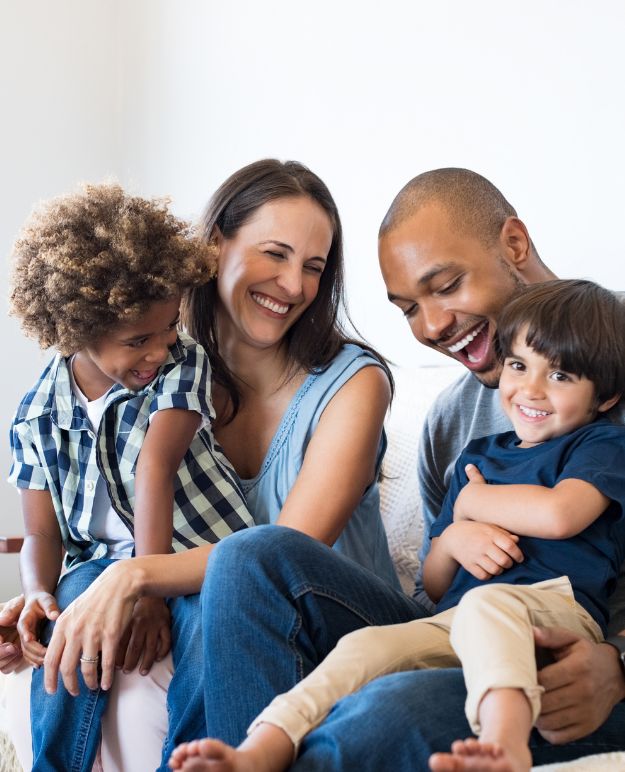 A couple laughing and playing with their kids on their living room couch.