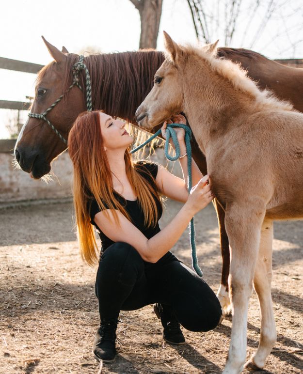 A young person is thrilled to embrace a foal under the watchful eye of the mare in a sunny paddock.