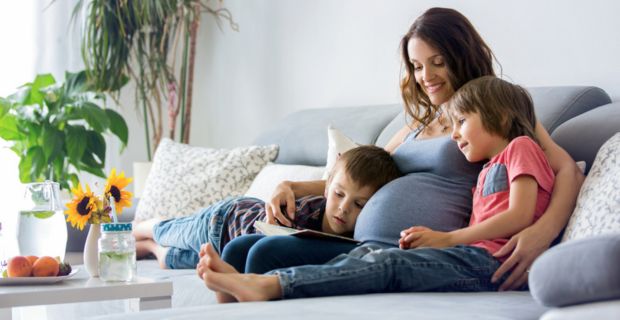 A pregnant parent snuggling with her two kids while reading to them on the couch.
