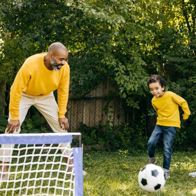 A Black father playing soccer with his son in the backyard.