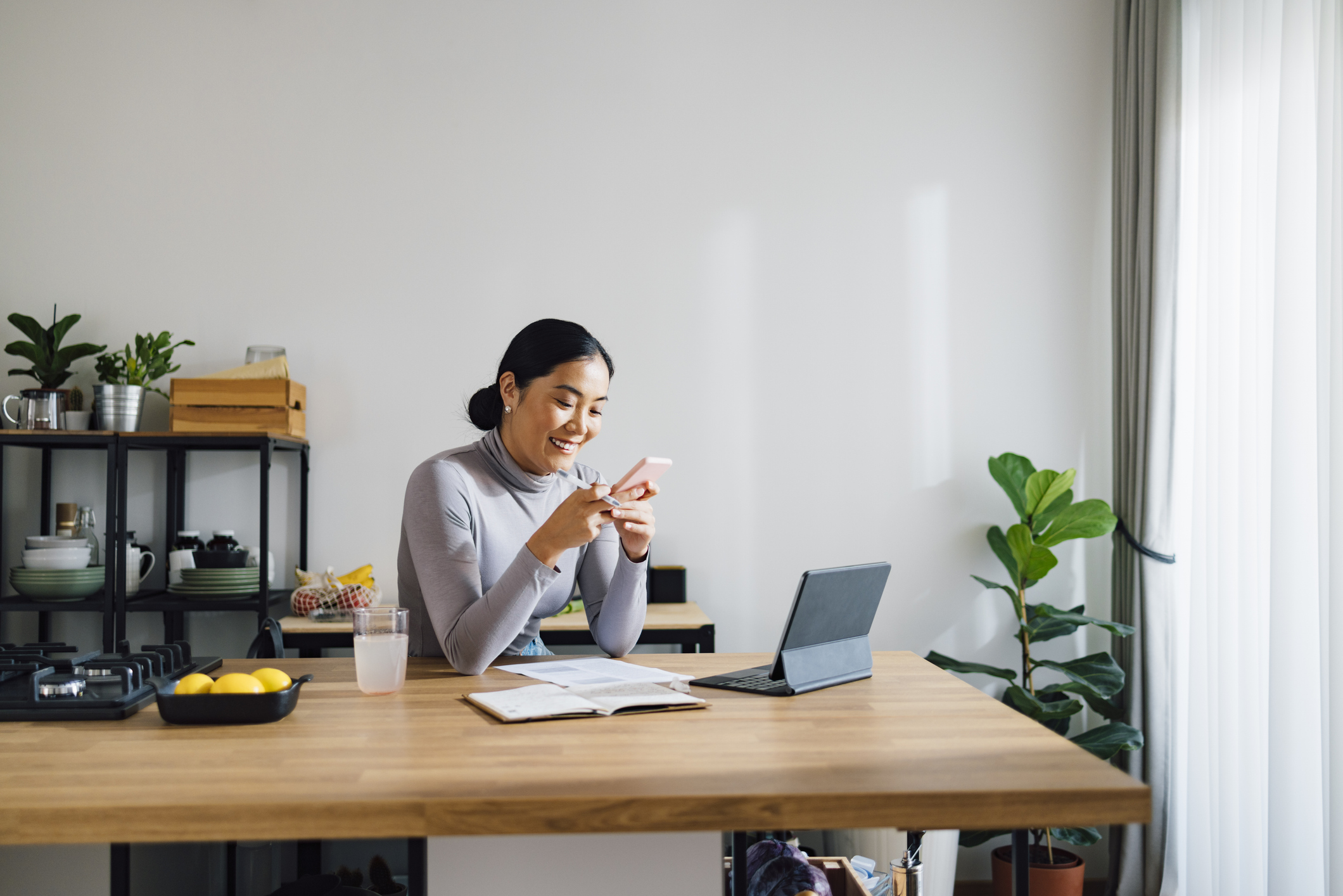 Woman sitting at kitchen table with laptop and document smiling at phone