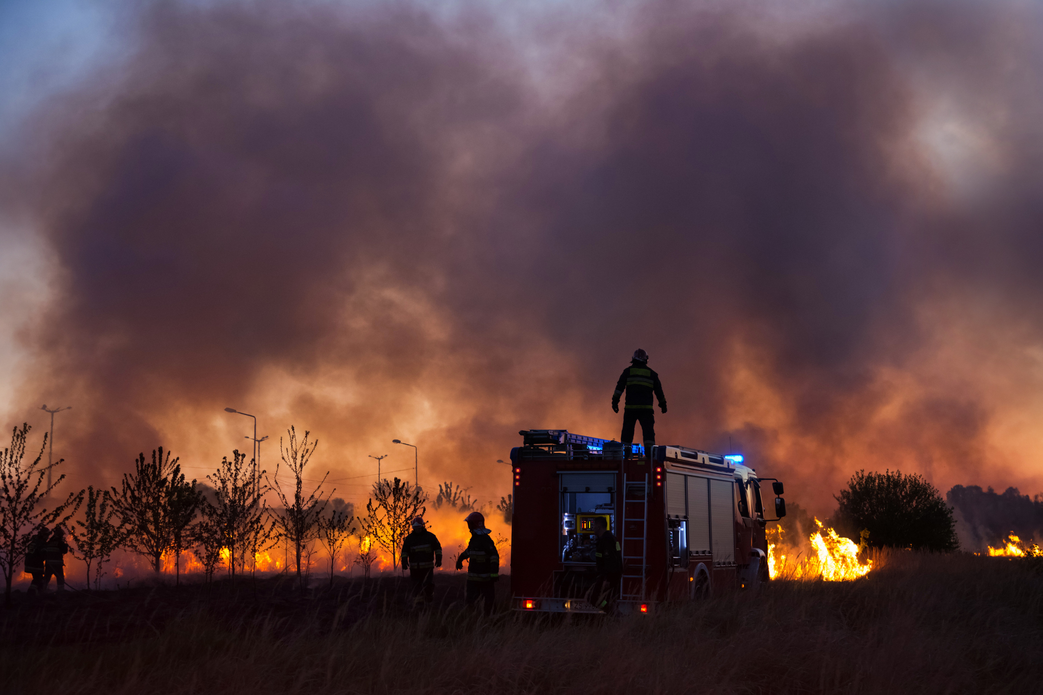 Firefighters standing around firetruck with wildfires and smoke-filled sky in background