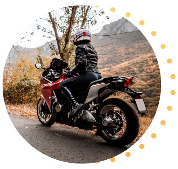 A motorcyclist in full gear sits on a red sports bike beside a road, framed by a scenic autumn mountain landscape.