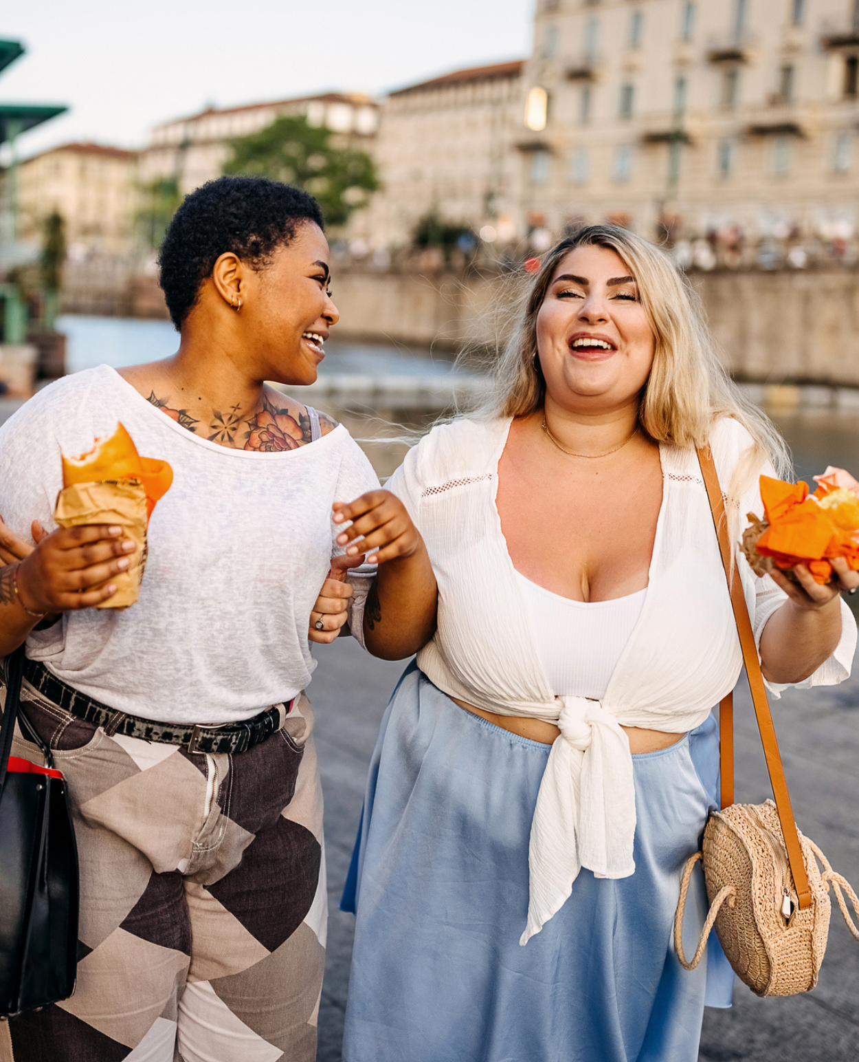 Two friends walk along the canal, laughing and enjoying local delicacies while on vacation. 