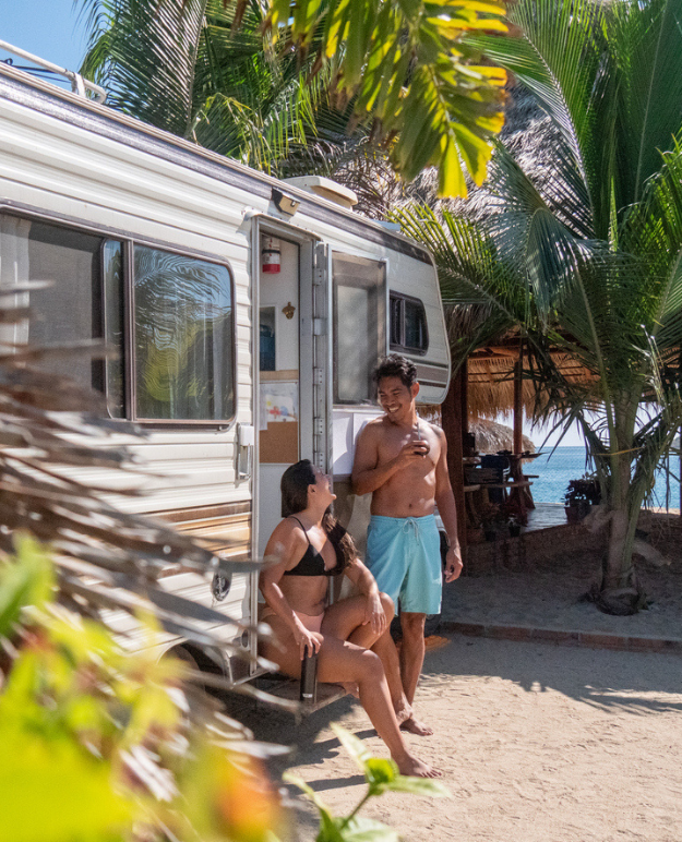 A tanned couple relaxes on the porch of their RV, parked on a sunny beach in Mexico.