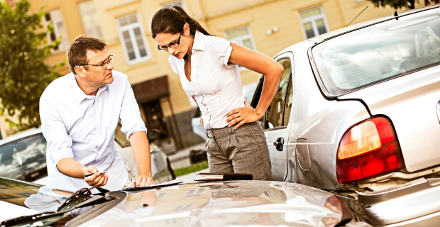 Two drivers looking frustrated while filling out an accident report after a staged collision in a parking lot.