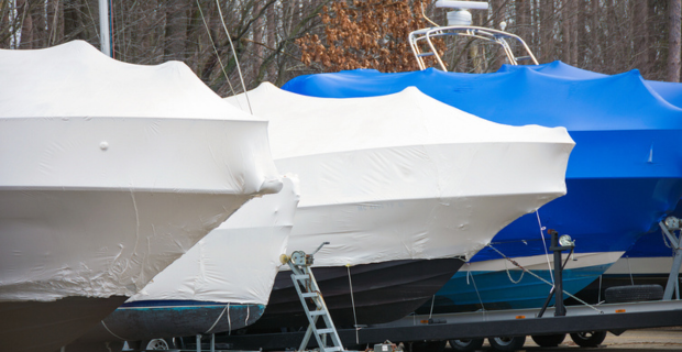 Side view of five boats with winter covers parked side-by-side on boat trailers for winter storage in front of a forest. 