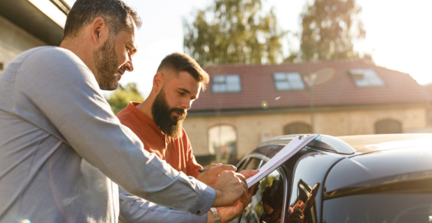 Two people stand beside a car discussing documents, illustrating the process of buying cars in Canada and navigating car prices.
