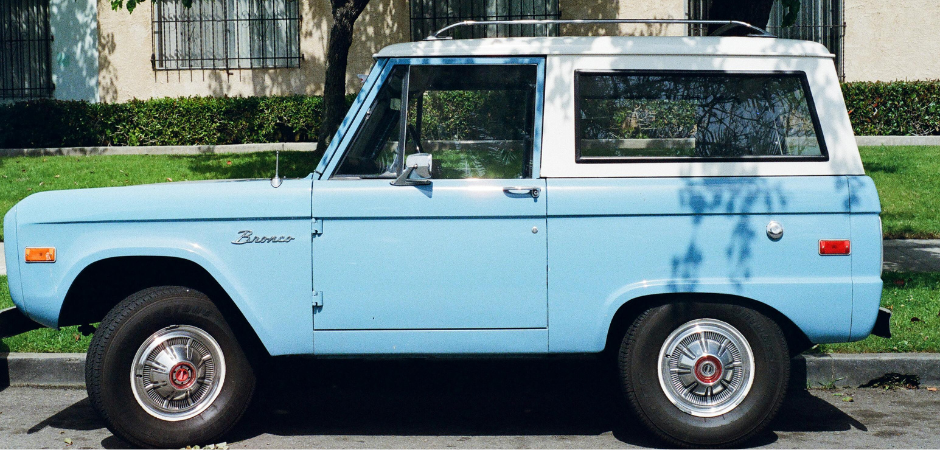 A light blue classic Ford Bronco two-door SUV from the early generation, parked on a street and viewed from the side profile.