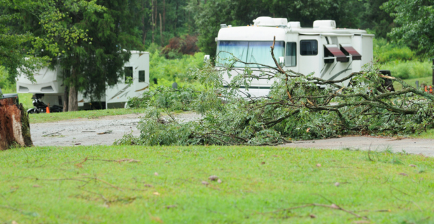 Two white RV’s can be seen parked in a campground. There are many branches seen scattered around the ground showing the aftereffects of a storm. There are many trees in the background.
