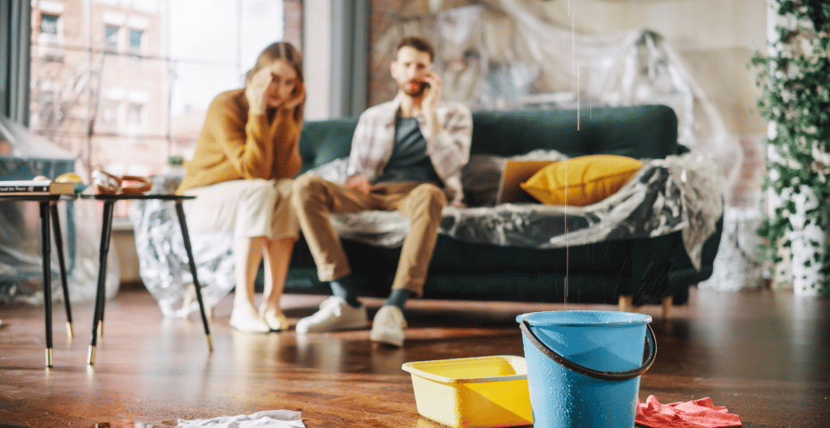 Water damage in a living room with buckets on the floor and furniture covered in plastic to catch leaks.