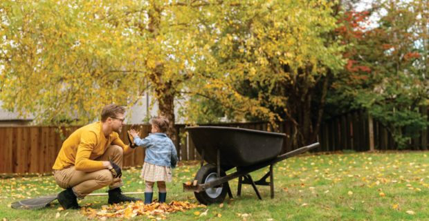 A person and a child in a yard with autumn leaves beside a wheelbarrow.