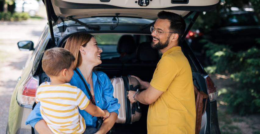 A happy family of three is seen smiling and chatting while packing their car trunk for a family road trip. 