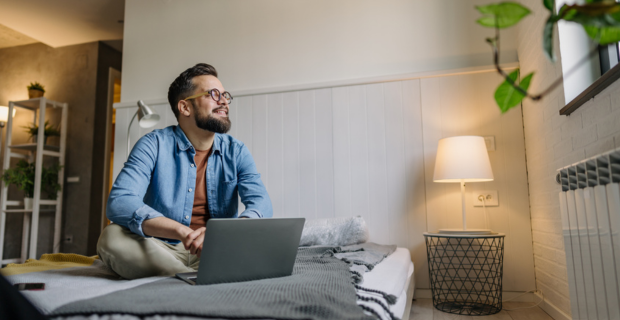 Young man in his apartment researching renters insurance on his laptop while sitting comfortably on his bed in a cozy, well-lit bedroom.