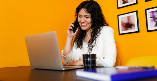 Happy adult woman talking on the phone in front of her laptop sitting at a desk in her office with orange walls and some pictures hung up behind her.