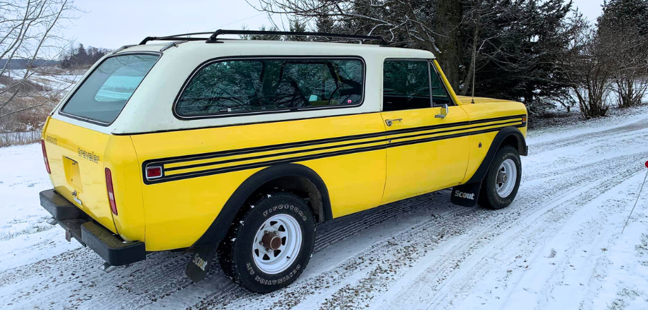 A bright yellow International Harvester Scout II SUV from the 1970s parked on a snowy gravel driveway, viewed from the rear-left angle.