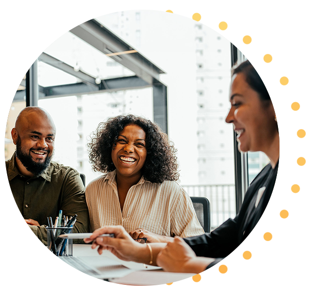 Smiling man and woman sitting at desk in office setting facing woman looking at a laptop