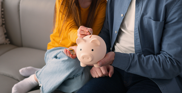 A young girl, with her father by her side, drops a coin into a pig‑shaped money bank.
