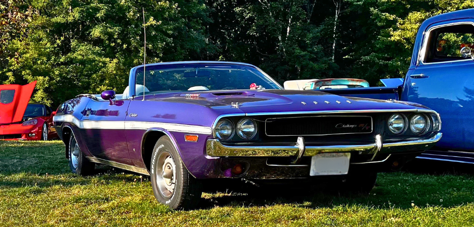 A classic purple Dodge Challenger R/T convertible parked on grass at an outdoor car show, photographed from the front-left angle.