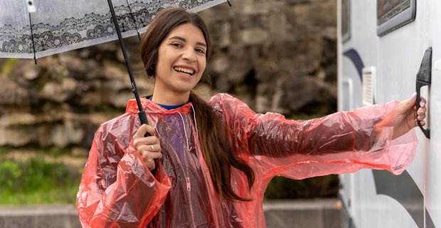 A woman seen smiling with an umbrella and rain poncho about to open the door to her motorhome.