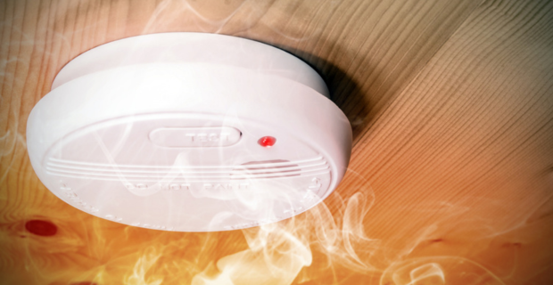 An image of a smoke detector attached to a wood paneled roof with smoke rising to it. 