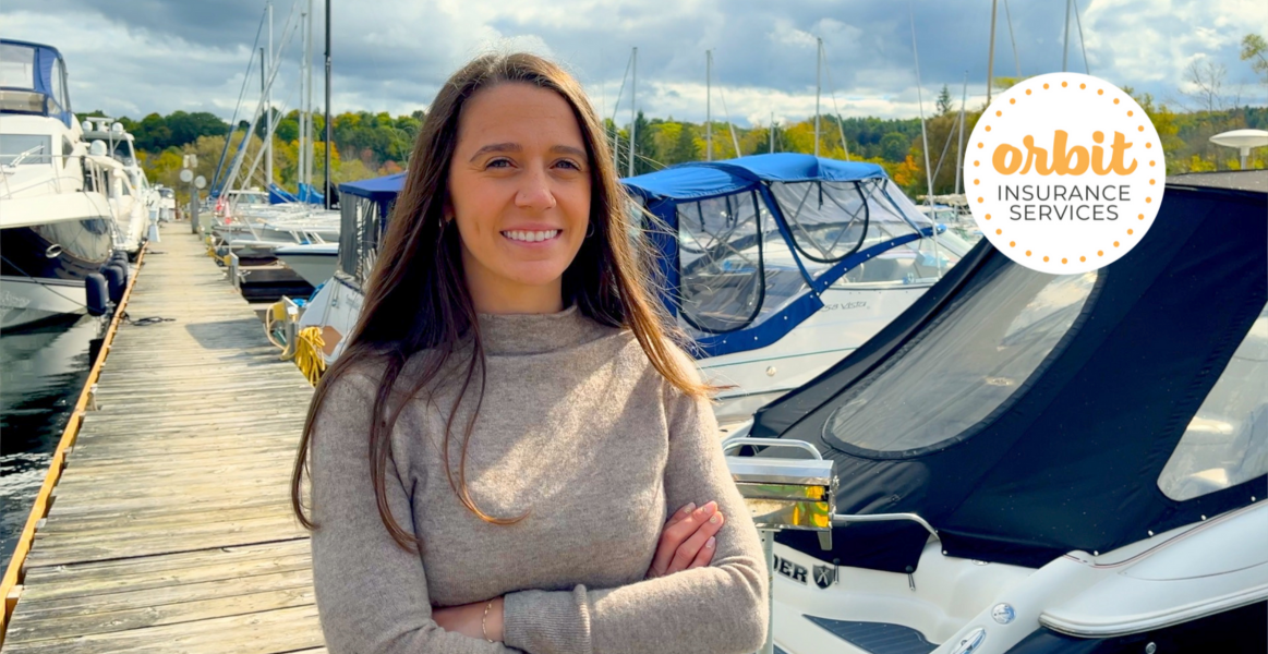 A person with long hair stands on a dock talking about marine insurance. They’re smiling and wearing a beige sweater while several boats are docked behind them. 