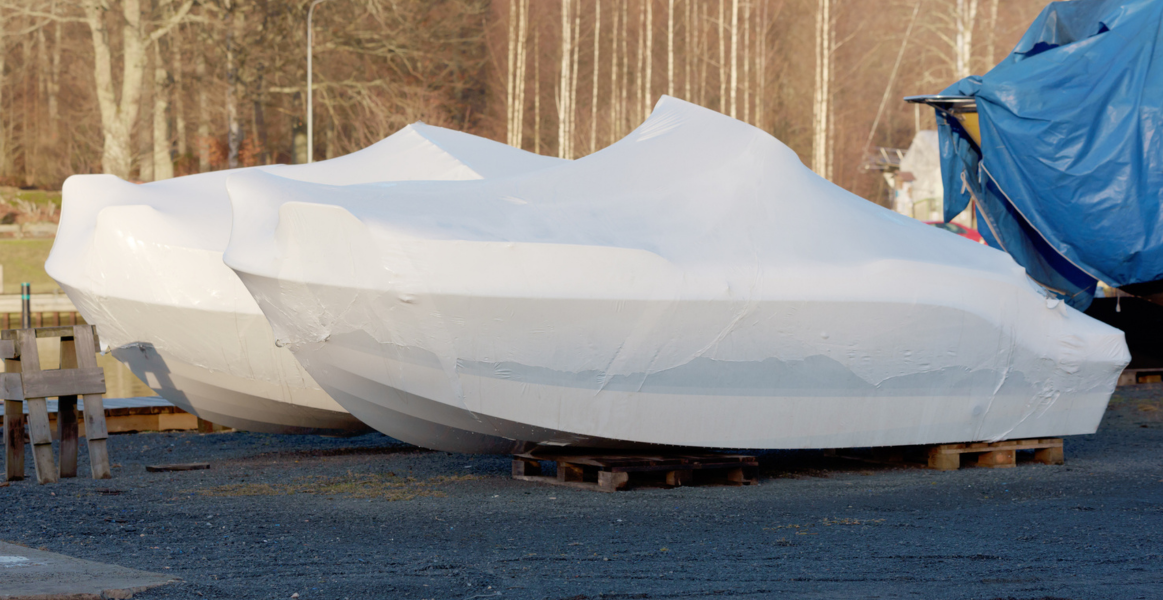 Two boats covered in white shrink wrap for winter storage, placed on wooden pallets outdoors near trees and a blue tarp-covered structure.