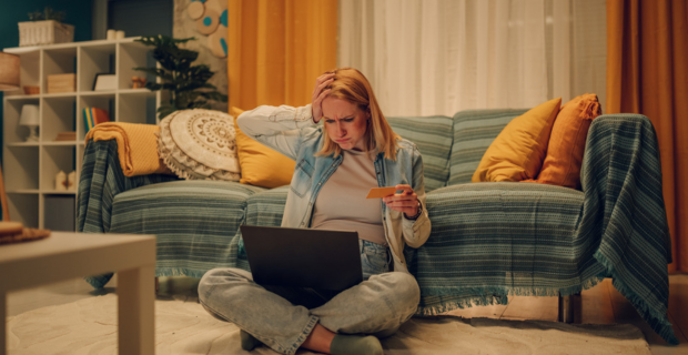 A person sits on the floor of their living room, holding a credit card and looking worriedly at their laptop, suggesting a possible encounter with an online shopping scam.