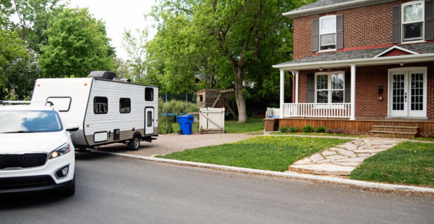 A white truck is backing a new white trailer into a driveway. There is a red brick home next to the truck and trailer. The yard has lush green trees and a play fort. There is a green recycling bin to the left of the trailer.