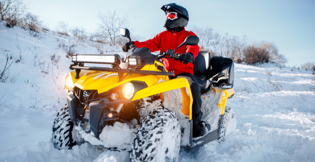 A person in a red jacket and black helmet rides a bright yellow ATV through deep snow on a sunny winter day with ATV insurance.