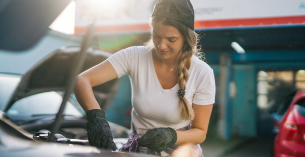 A young woman looking under the hood of a vehicle performing some classic car spring maintenance to get it ready to drive.