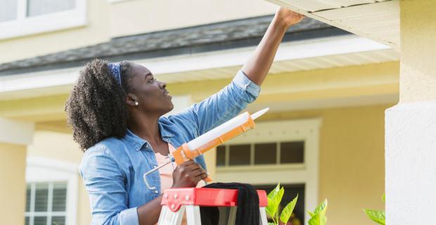 A woman is seen on a ladder next to her house on a sunny day with a caulking gun sealing here roof to protect against spring flooding.