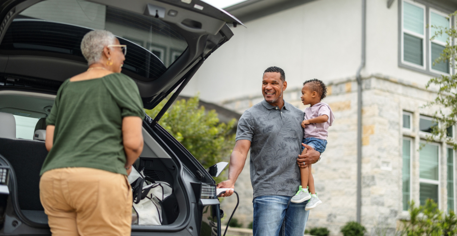 A person with short gray hair is unpacking their EV while another person holds a child and charges the vehicle. The car is parked in a residential area, and they’re talking about usage-based insurance.   