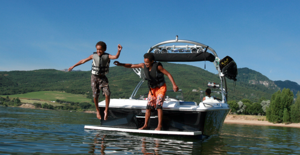 Two young boys seen jumping off a wake boat on a lake with life jackets on.  